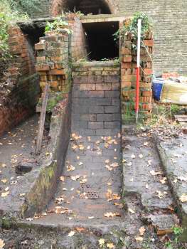 Brick channel with a low wall on the left and the remains of a wall on the right, leading up to a lip with a dark opening above it.
