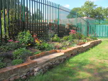 A raised flower bed is situated between a green paling fence and a grassy path.