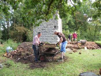 Two men seiving spoil into a wheelbarrow, with two more men and a the base of a stone chimney in the background.